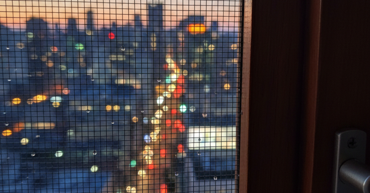 منظر ليلي من خلال شبك ناموس مثبت على نافذة خشبية يظهر أضواء المدينة والمباني في الأفق وقت الغروب.